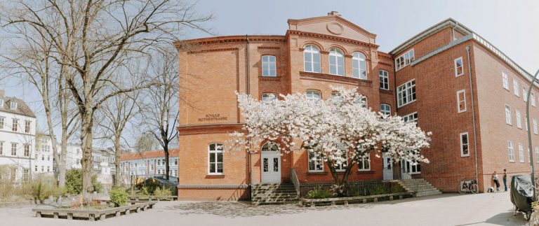 Das gesamte Gebäude wurde hier als Panoramafoto aufgenommen. Vor dem Backsteingebäuder der Schule blüht weiß eine große Zierkirsche. Diese steht rechts vor dem Haupteingang der Schule.